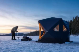 Insulated pop-up ice fishing shelter on a frozen Canadian lake with angler setting an ice anchor beside sled and rods