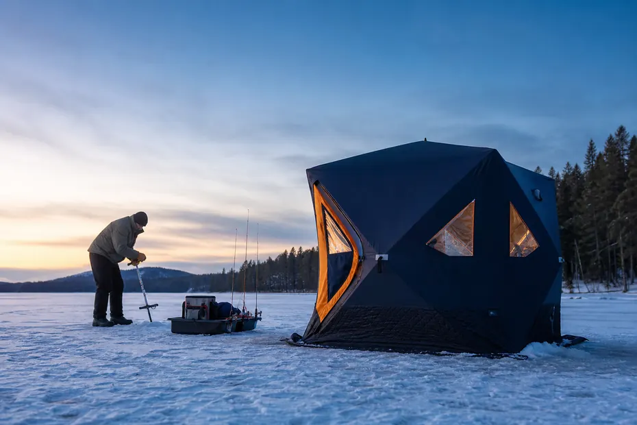Insulated pop-up ice fishing shelter on a frozen Canadian lake with angler setting an ice anchor beside sled and rods