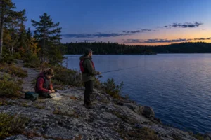 Two adults wearing PFDs fish and prepare tackle on a broad rocky lake shore at blue hour, with a headlamp directed towards their hands.